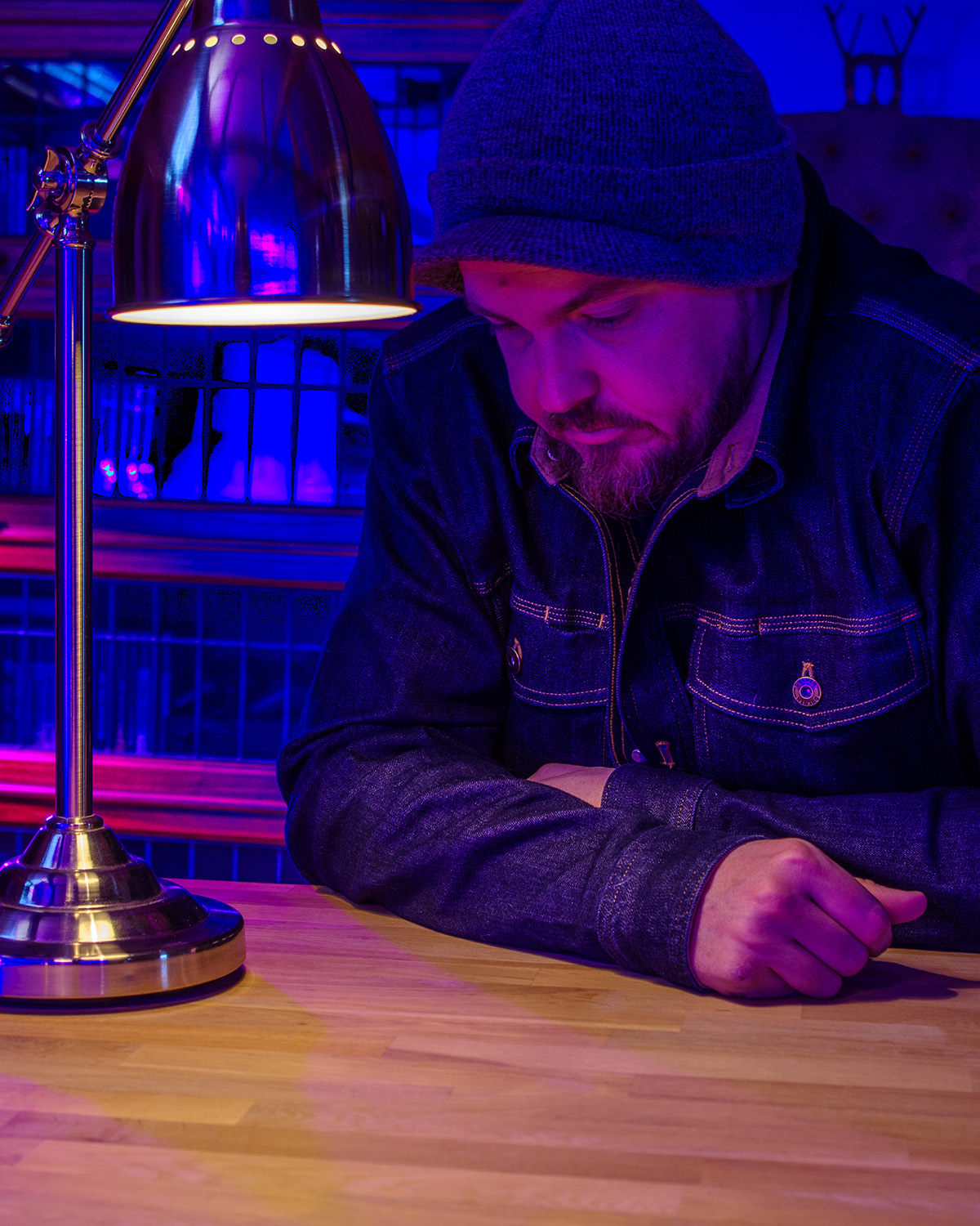 Sean seated at a wooden desk under a single lamp, head slightly bowed in concentration, blue and purple light filling the room. The image carries the current mode of the work — sustained, private, and deliberate engagement with the questions that now occupy the center of the practice.