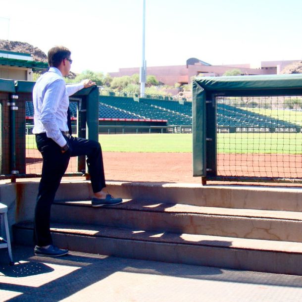 Sean in formal attire standing in a dugout, looking out over an empty professional baseball stadium in Arizona. The image anchors the operational years inside baseball — the quiet responsibility of maintaining the conditions under which the game could be played.