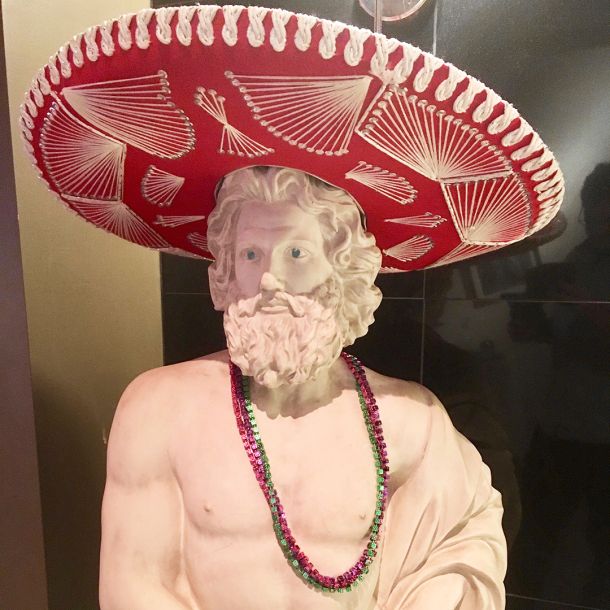Classical Greek statue of a philosopher. It's wearing a bright red sombrero and mardi gras party beads.