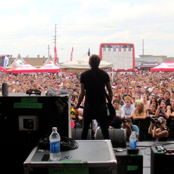 Looking out from the stage at a packed Warped Tour crowd under summer sky. The image carries the scale and logistics of those summers — radio, promotions, and daily operations across dozens of cities so the performances could take place.