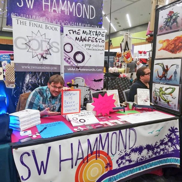 Sean at a convention table with banners for The Final Book: Gods and The Mixtape Manifesto, books stacked and handwritten signs on display. The image holds the public-facing author years — the practice of putting the work in front of people and standing behind it.